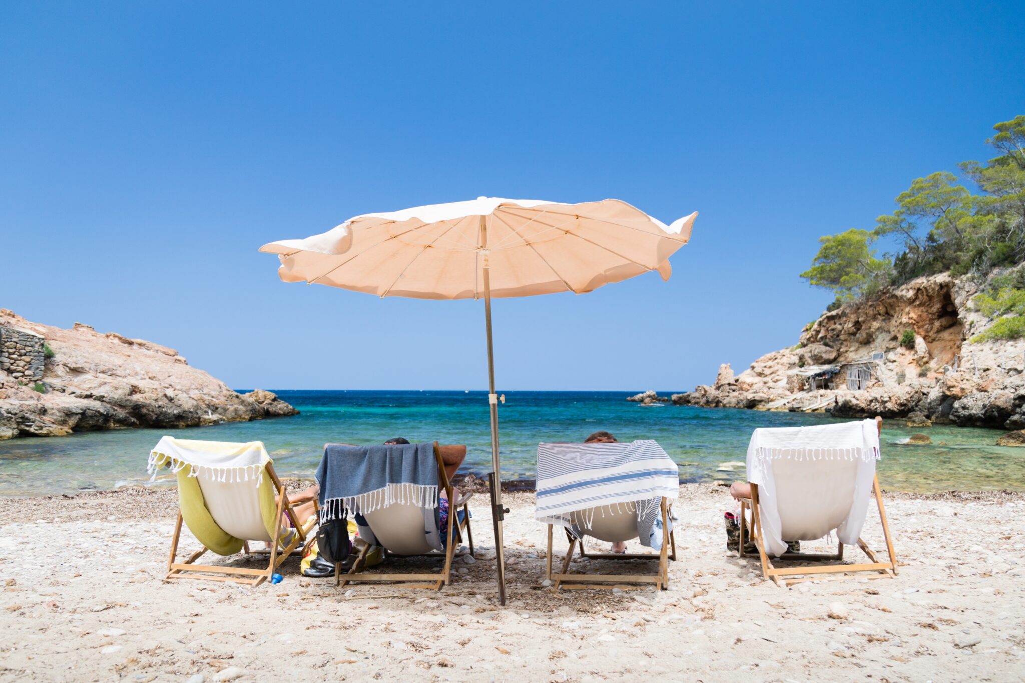 A shot of four  people in sunbeds on the beach, sunbeds covered with towels, placed next to each other, around a beach umbrella